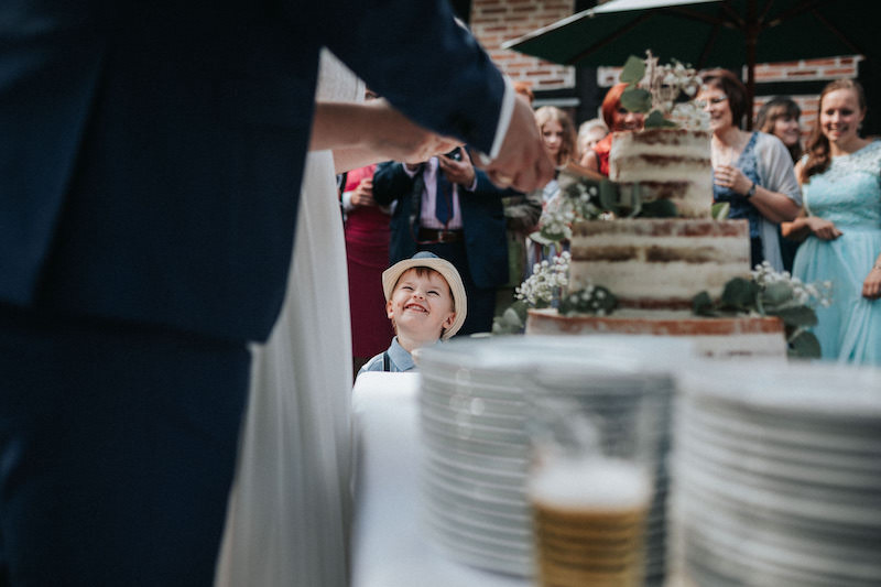 Junge lacht und schaut dem Brautpaar beim Anschnitt der Hochzeitstorte zu.